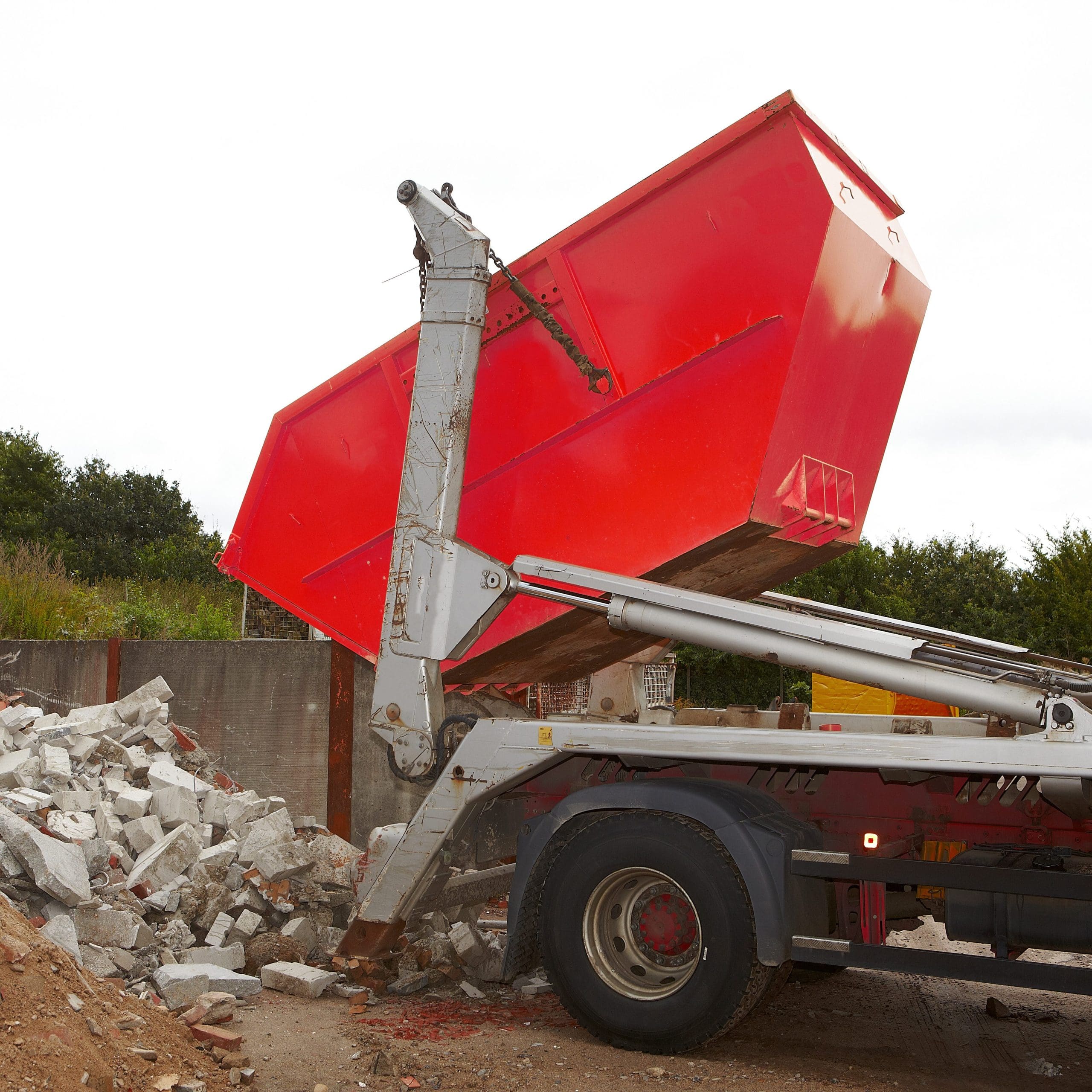Red construction skip being unloaded from a truck onto a pile of rubble and bricks at a construction site. Trees are visible in the background.