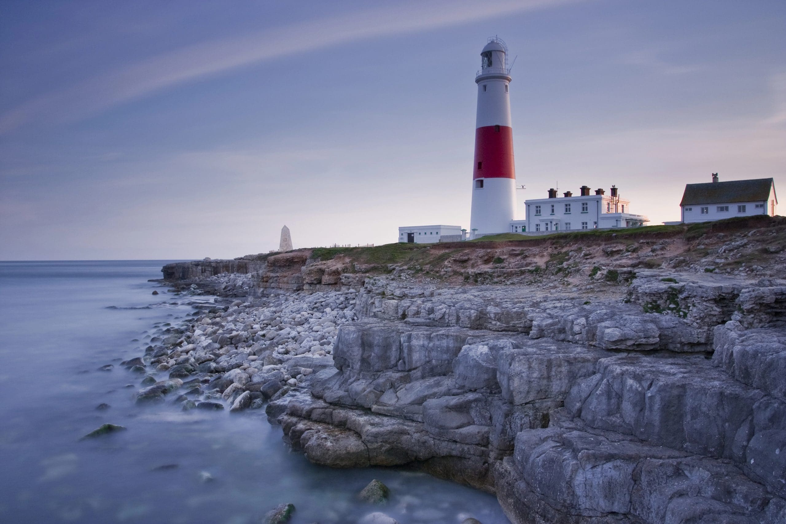 A lighthouse with a red and white tower stands on a rocky coastline at dusk. The calm sea is on the left, and buildings are adjacent to the lighthouse on the right. The sky is a soft gradient of blue and pink.
